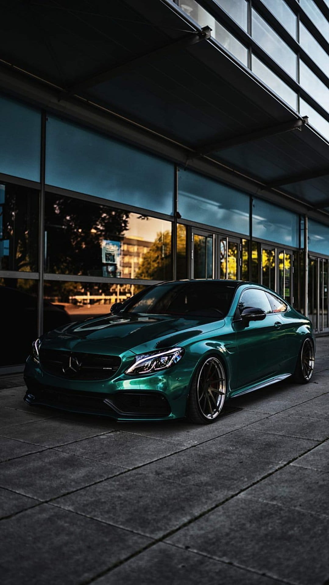 Emerald green Mercedes coupe parked on gray concrete tiles before a contemporary glass and steel building.
