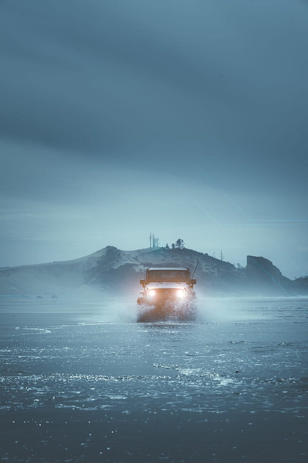 Jeep Wrangler with glowing headlights drives through splashing ocean waves on a misty, desolate coastal beach.