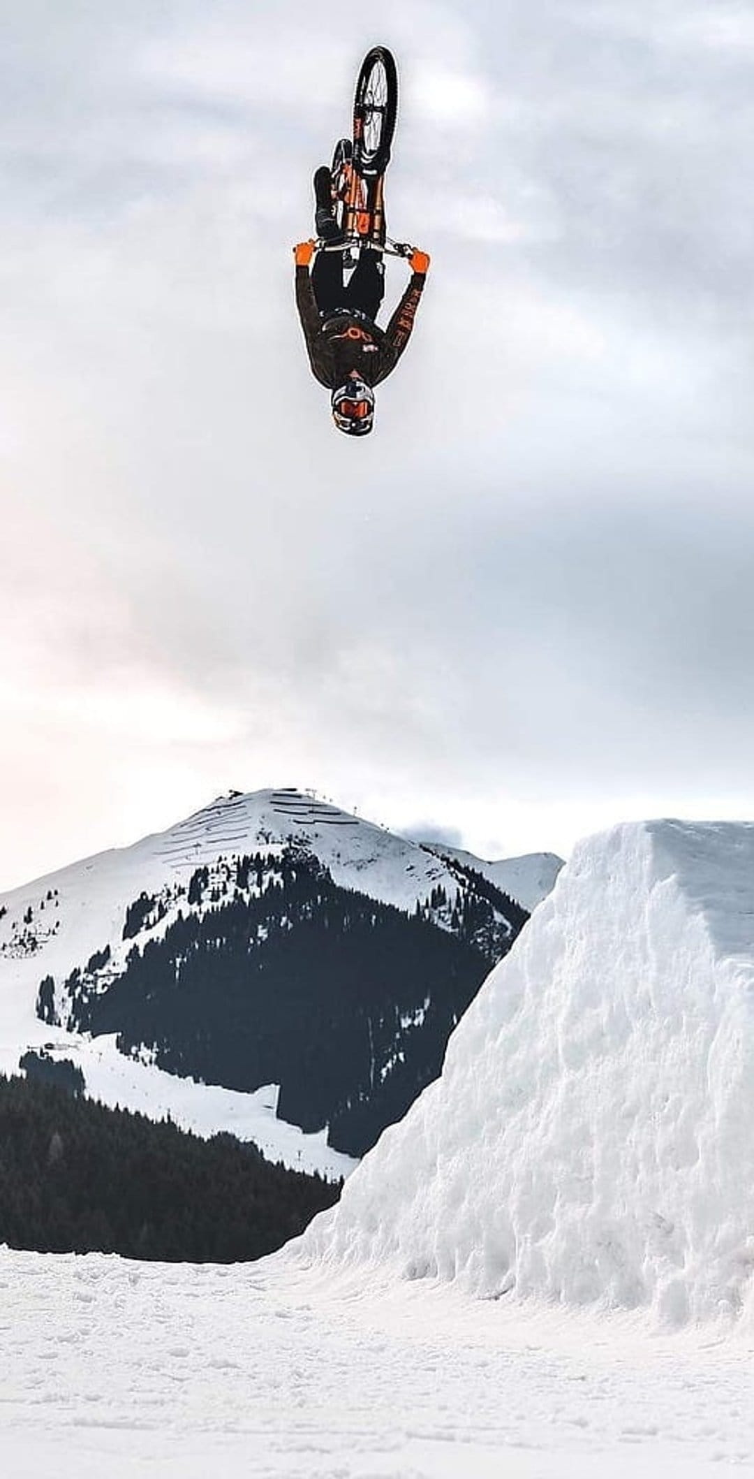 Mountain biker performs a backflip over a snowy ramp against a cloudy sky and pine trees in a winter landscape.