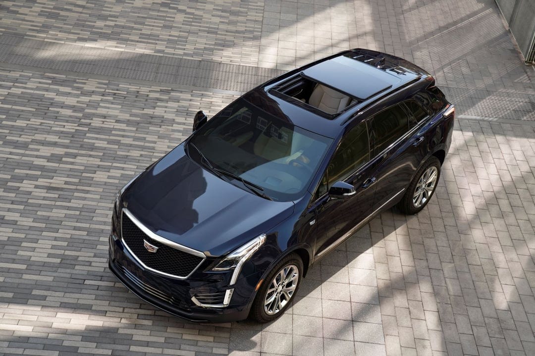 Dark blue Cadillac SUV from a high angle on a grey stone tiled plaza with an open sunroof and beige interior.