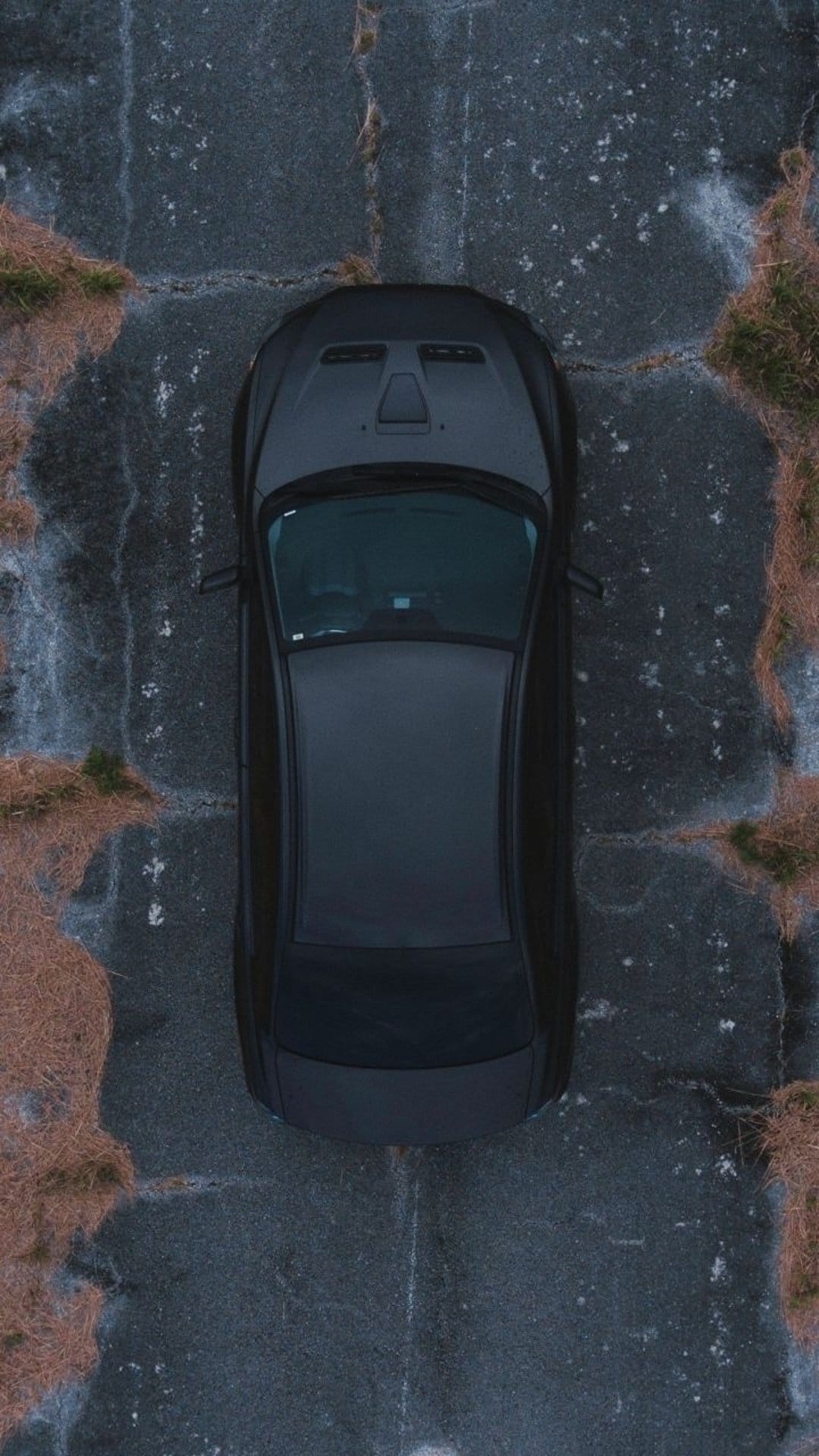 Matte black car viewed from above sitting on cracked grey asphalt surrounded by patches of dry yellow grass.