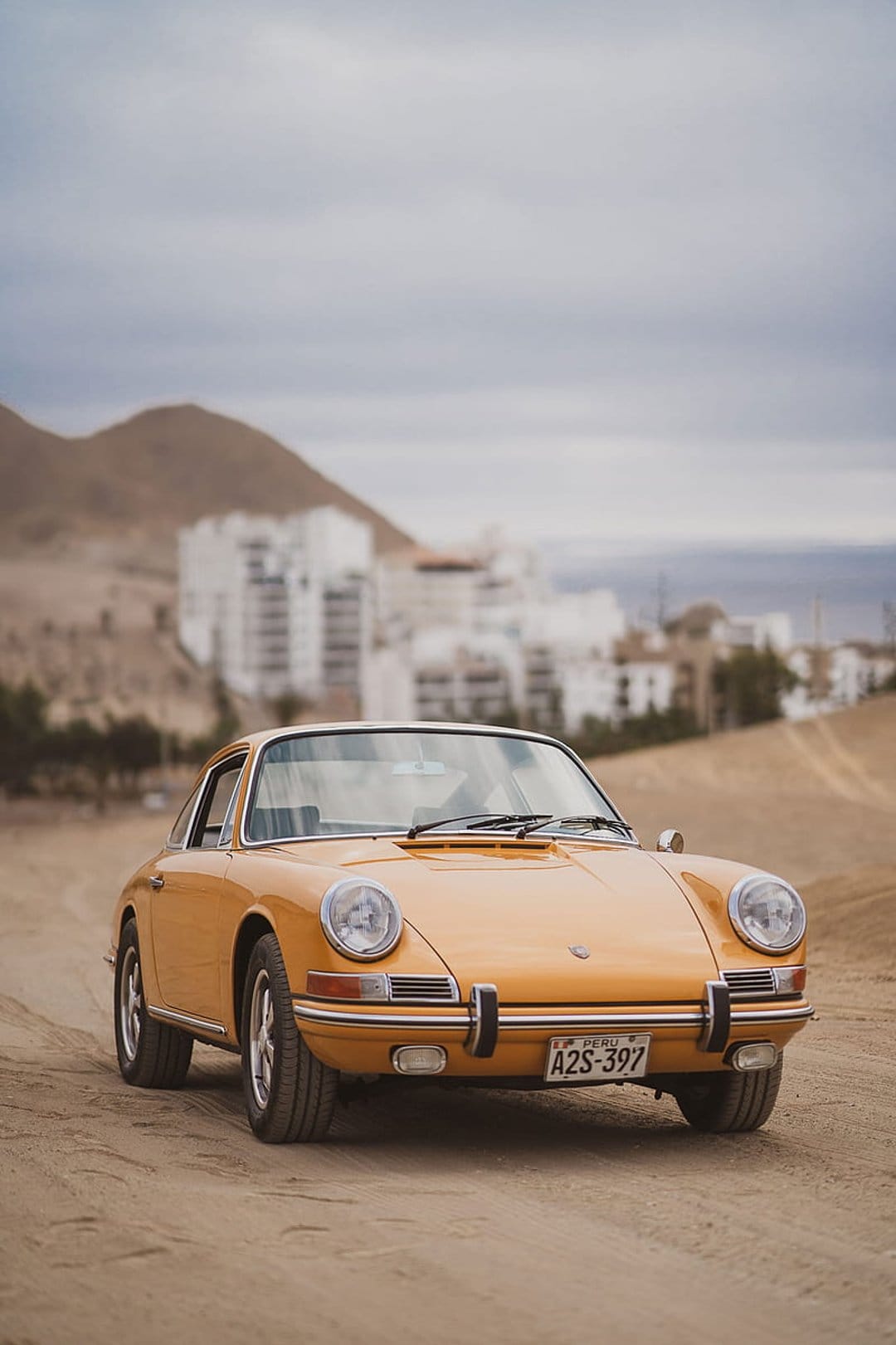Yellow vintage Porsche 911 parked on a dusty trail with rocky peaks and white buildings under a cloudy sky.