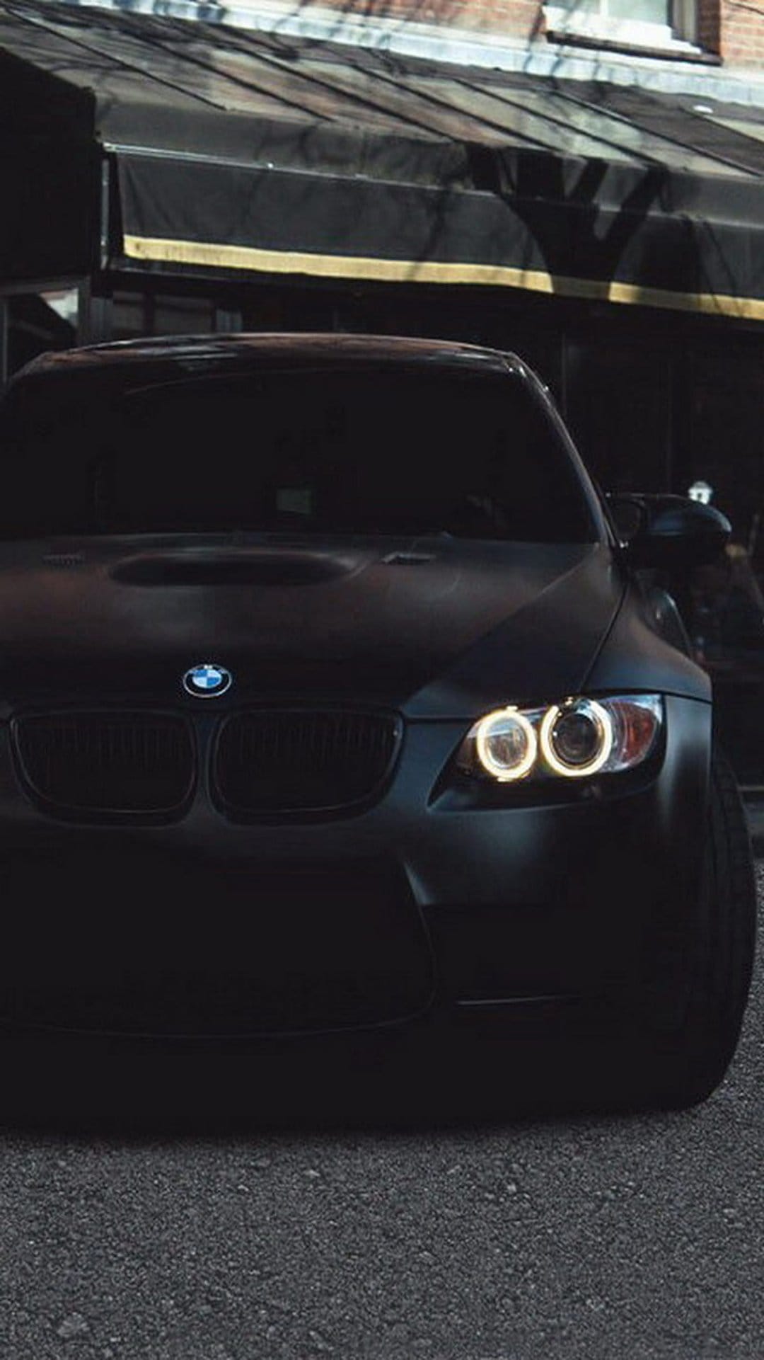 Matte black BMW M3 with illuminated angel eyes headlights parked on asphalt against a blurred brick building.