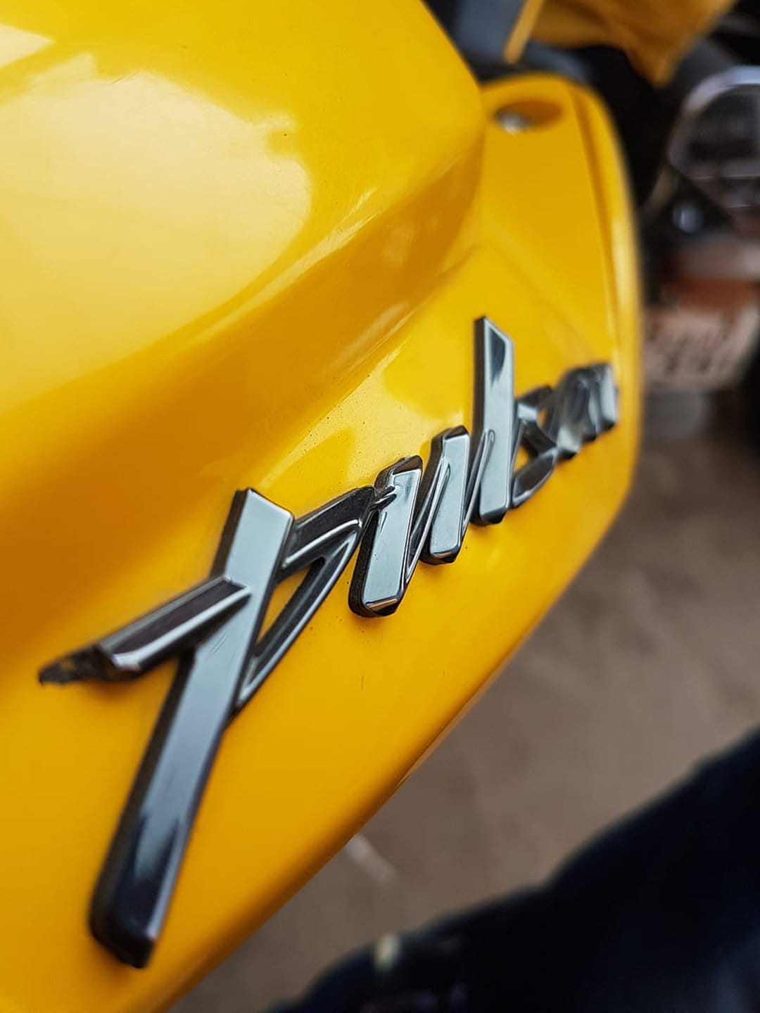 Pulsar chrome emblem on a bright yellow motorcycle body with a soft-focus background and metallic reflections.