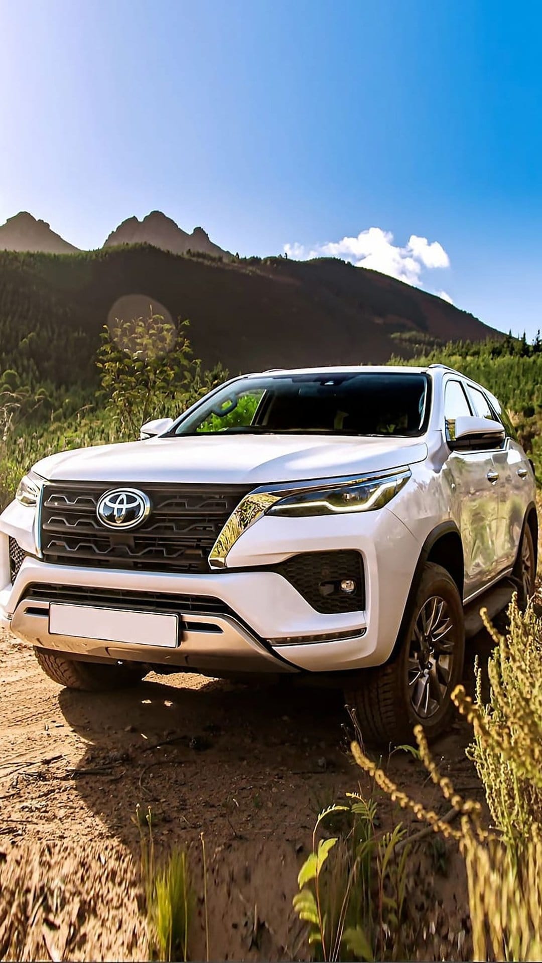 White Toyota Fortuner SUV driving on a narrow dirt path through lush green mountains under a clear blue sky.