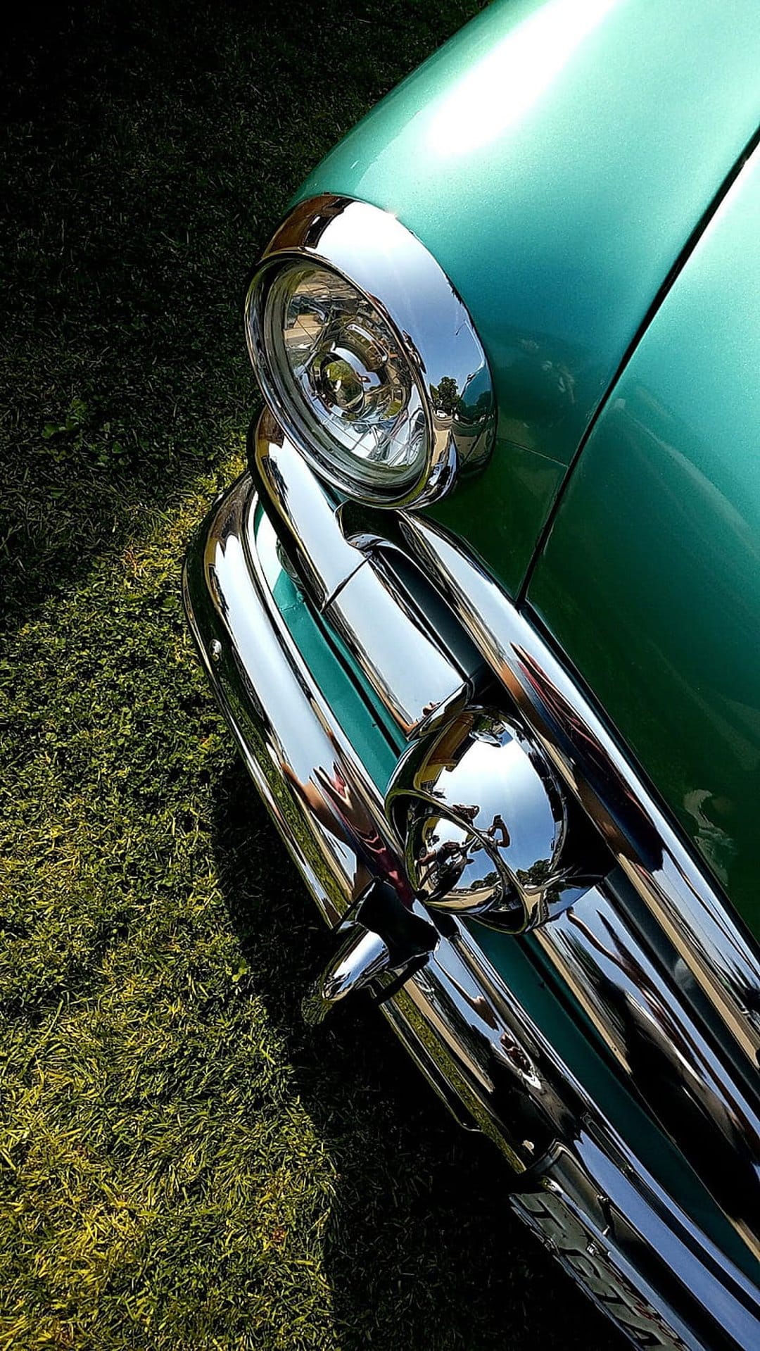 Teal vintage car headlight and reflective chrome bumper resting above vibrant green grass in a macro close-up shot.