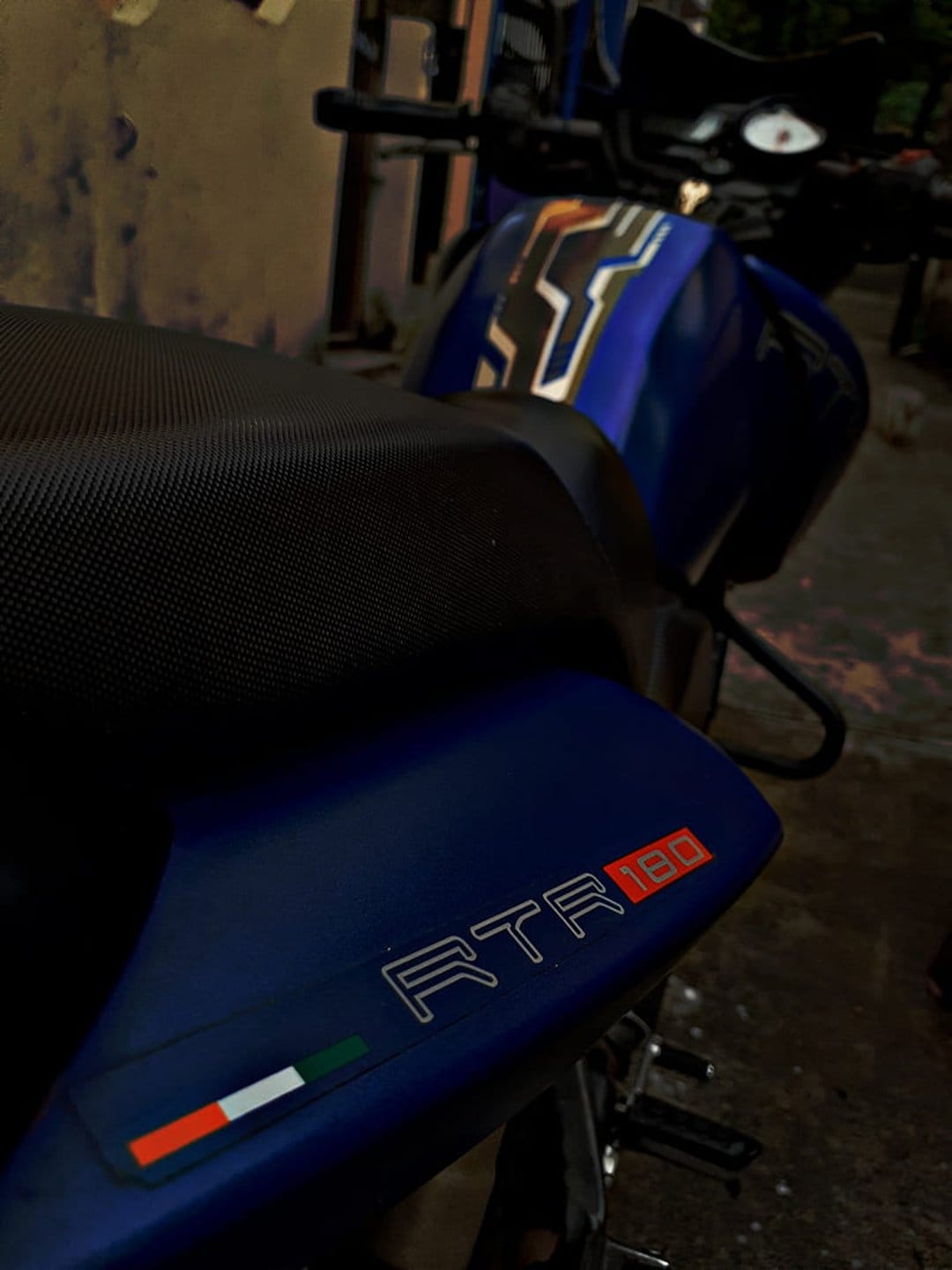 Blue TVS Apache RTR 180 motorcycle features a textured seat, RTR 180 badging, and a small Italian flag decal.