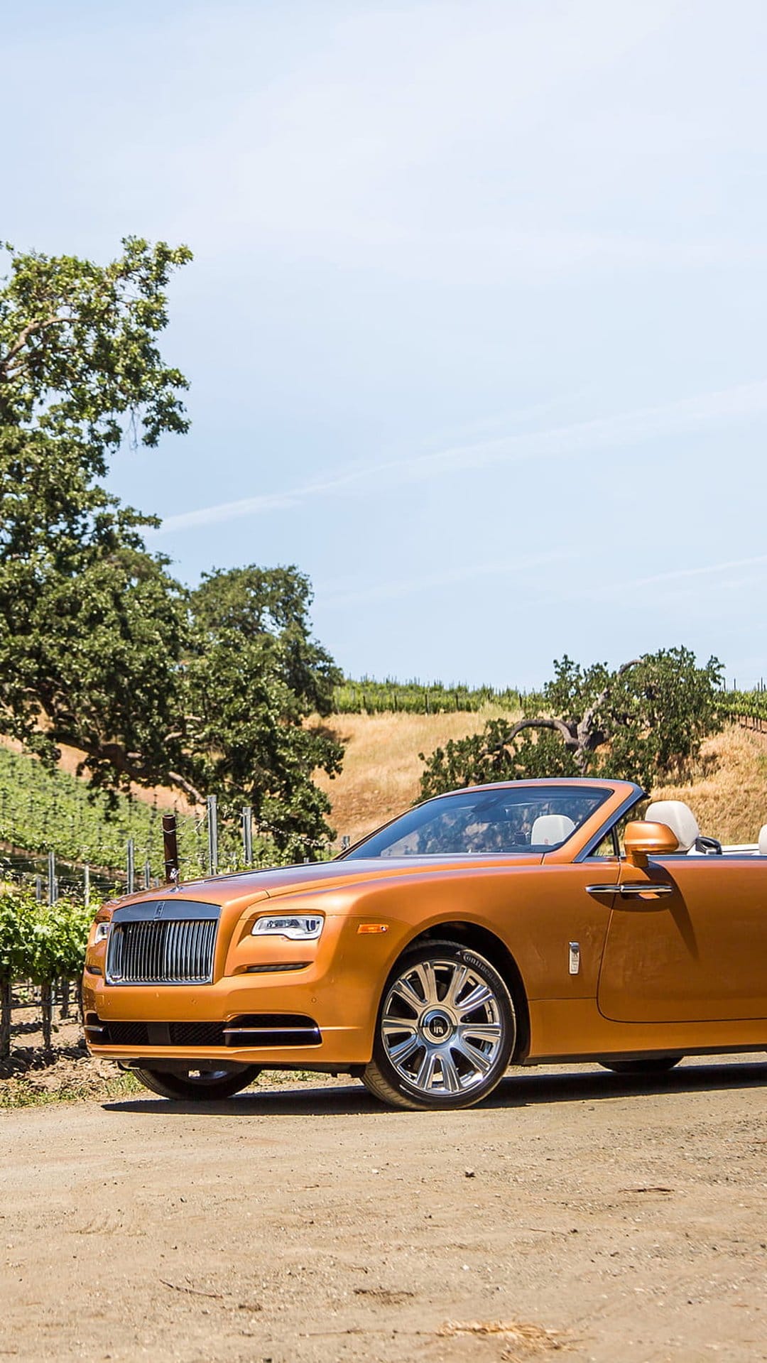 Orange Rolls-Royce Dawn convertible parked on a dirt road with vineyards and rolling hills under a blue sky.