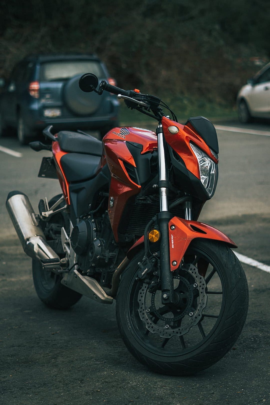 Orange Honda motorcycle parked on an asphalt road with visible disc brakes, exhaust pipe, and background cars.