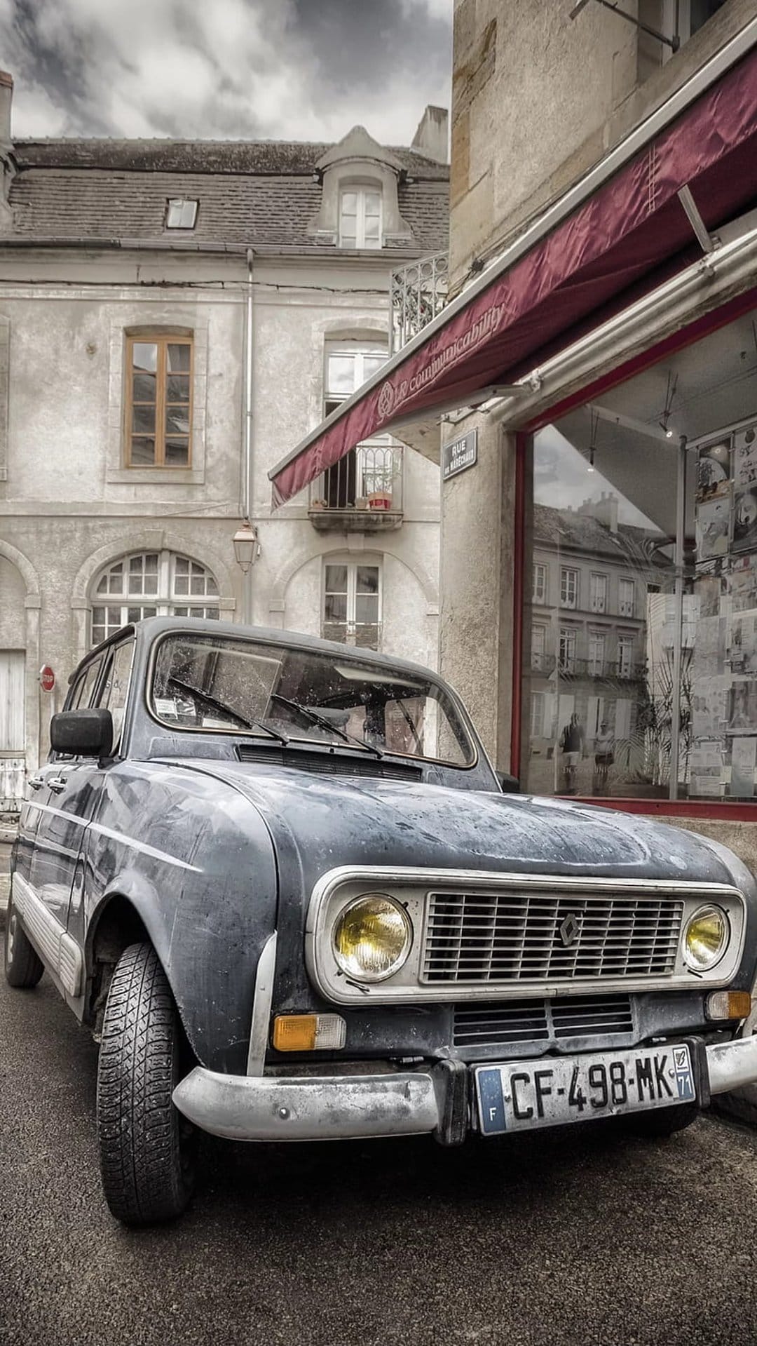 Dark blue Renault 4 parked on damp cobblestones in front of stone buildings with a red awning in France.