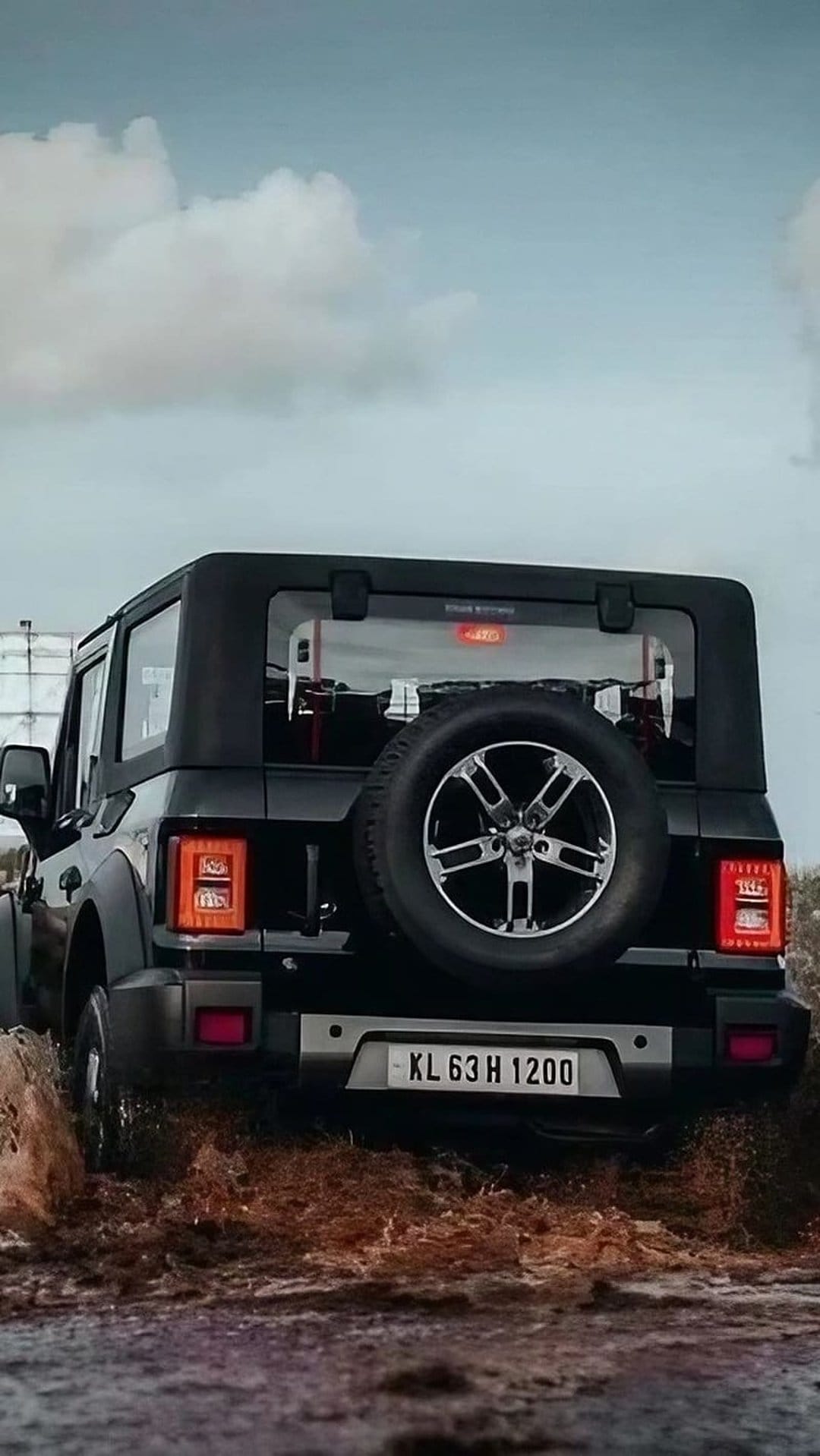 Black Mahindra Thar SUV rear view driving through a thick muddy track under a dramatic overcast gray sky.