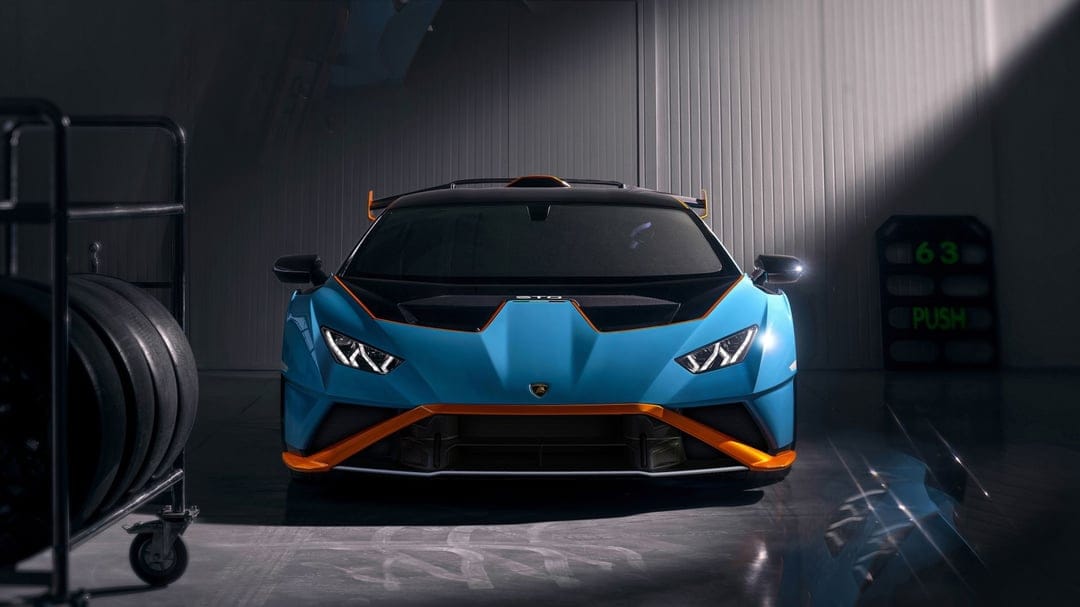Blue Lamborghini Huracan STO with orange trim in a dark garage under a spotlight beam with racing tires.