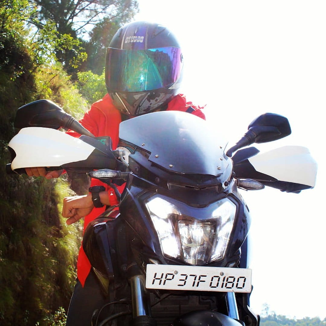 Motorcyclist in a red jacket and silver helmet sits on a black bike against a blurred green background.