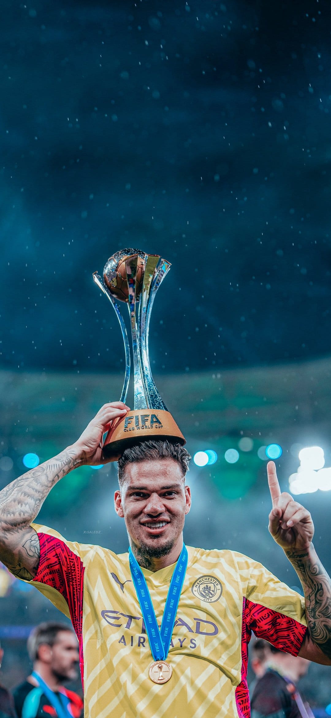 Manchester City goalie Ederson lifts a gold trophy wearing a yellow uniform and medal during a stadium celebration.