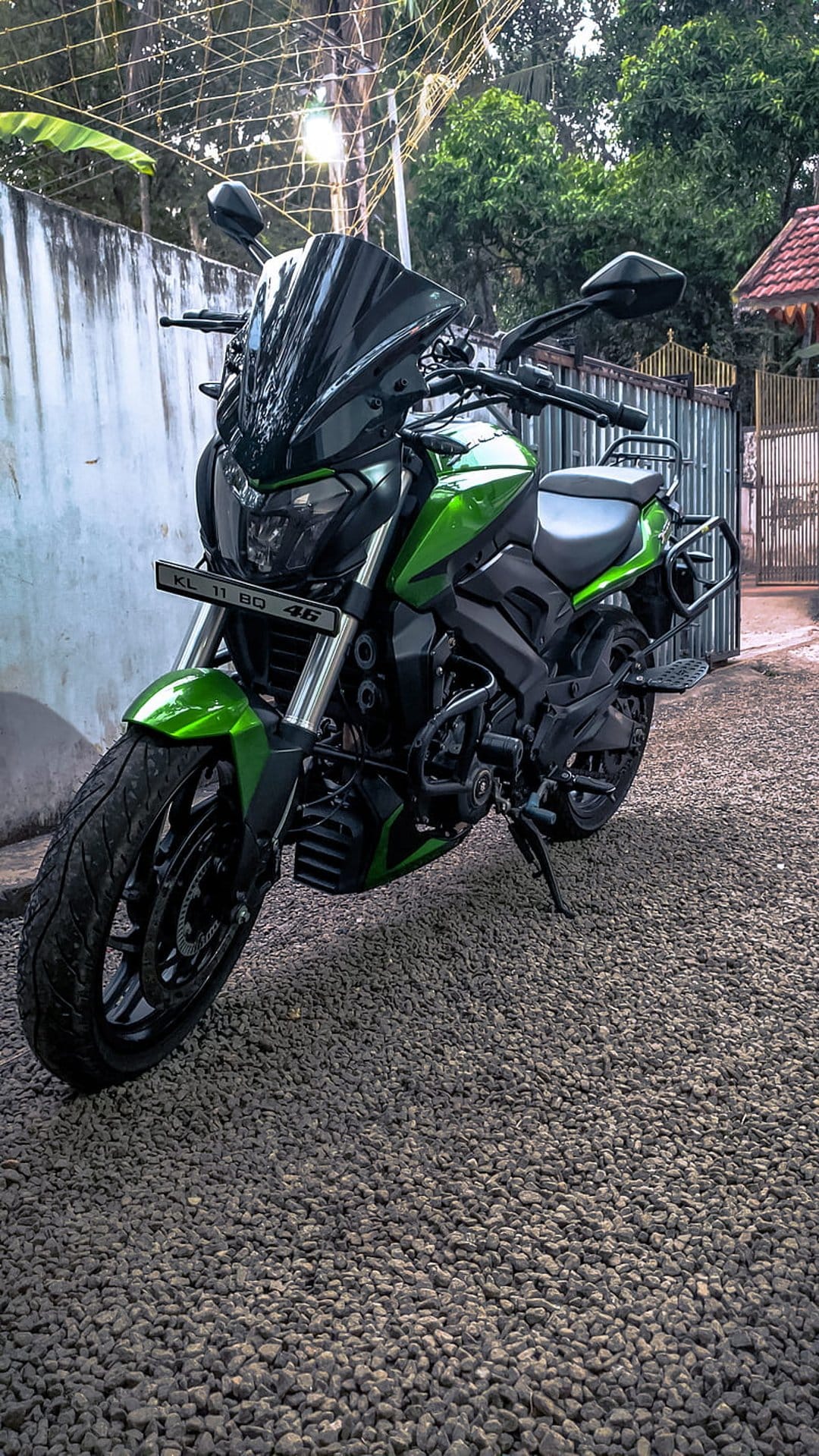 Green Bajaj Dominar motorcycle with black accents parked on a gravel road near lush green trees and a fence.