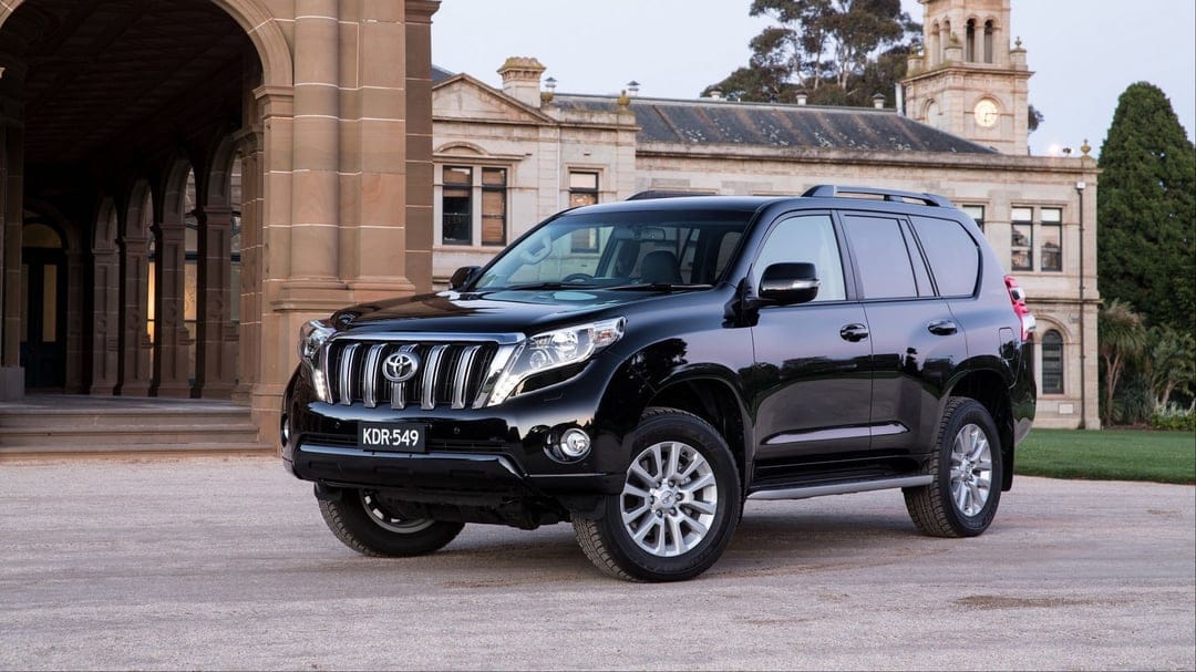 Black Toyota Land Cruiser Prado parked in front of a grand sandstone manor with a clock tower and arches.