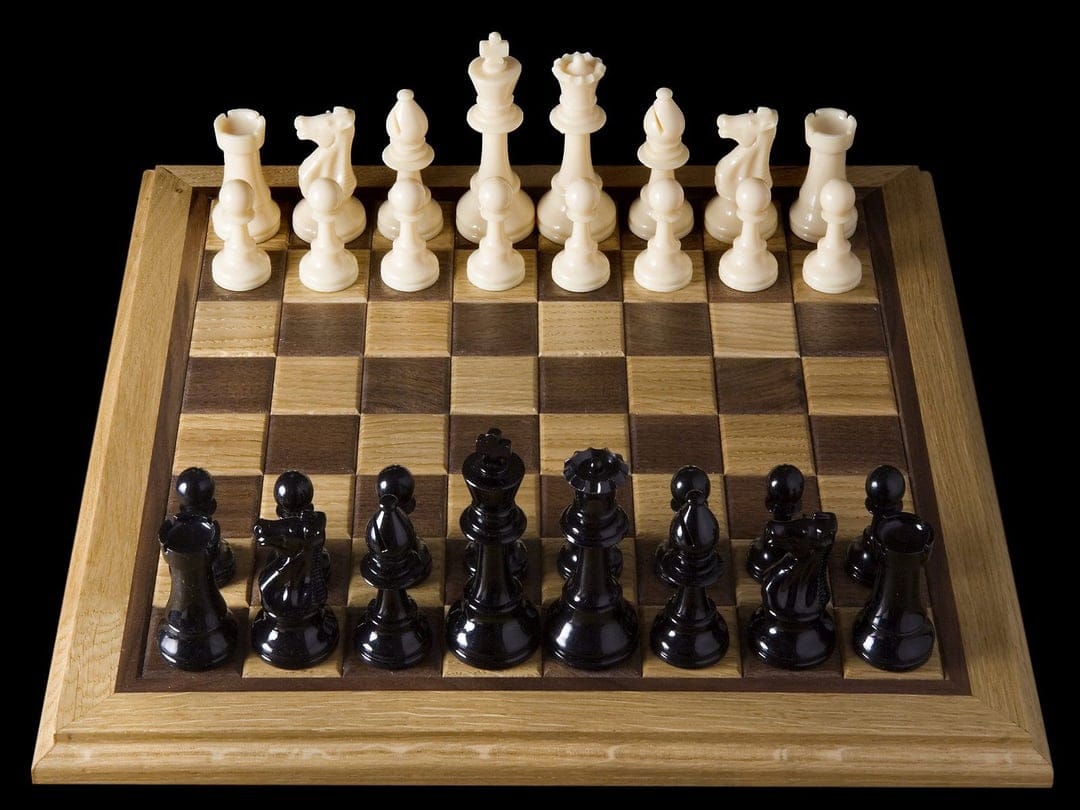 Black and white chess pieces arranged in starting formation on a wooden board against a dark background.