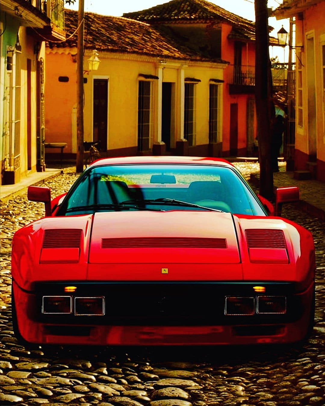 Red Ferrari 308 GTB parked on a cobblestone road beside colorful colonial houses during golden hour sunlight.