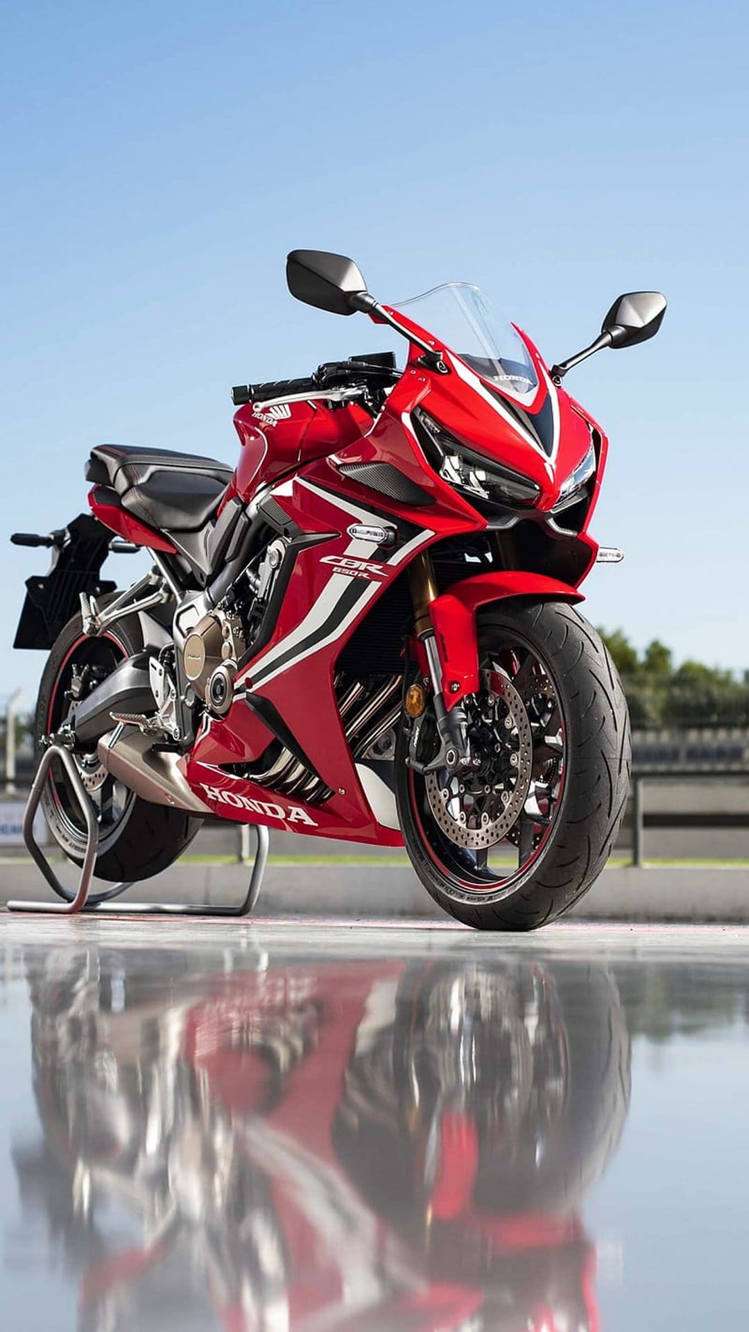 Red Honda CBR motorcycle sits on a stand reflecting onto a glossy ground under a clear blue sky.