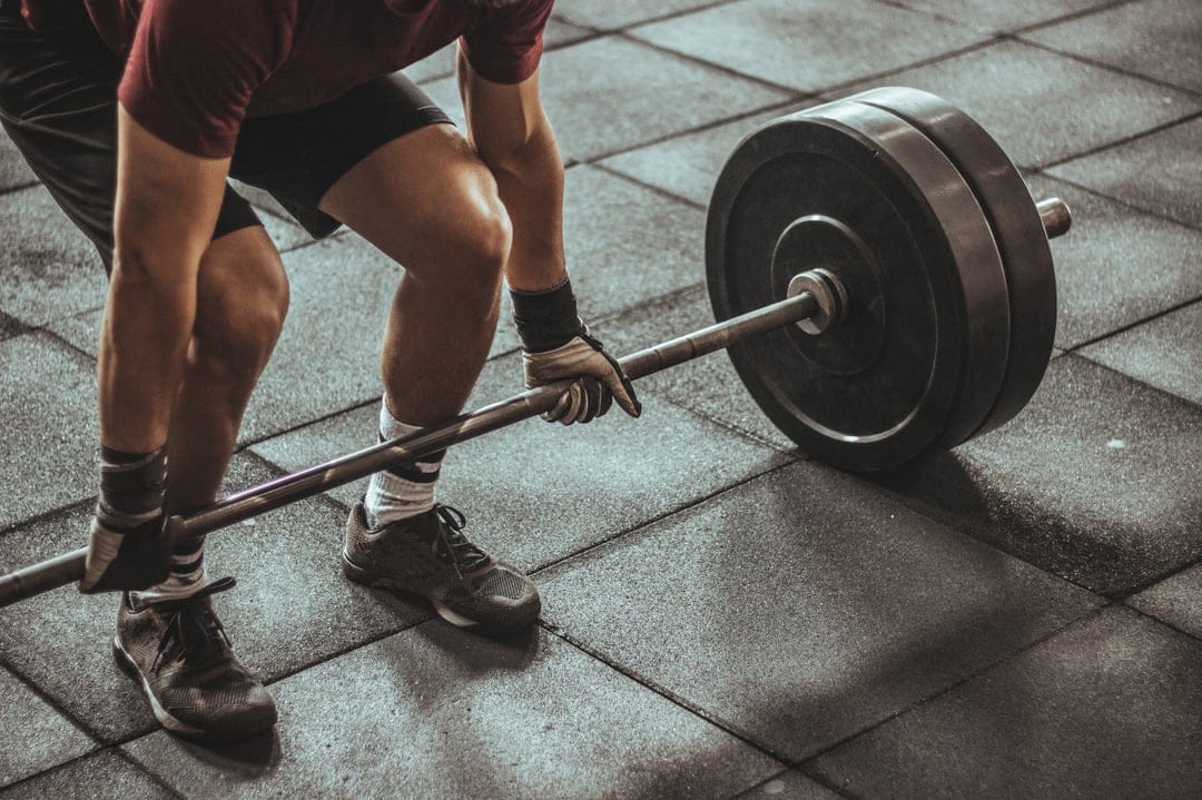 Muscular man wearing lifting gloves prepares to hoist a heavy barbell from a textured black rubber gym floor.