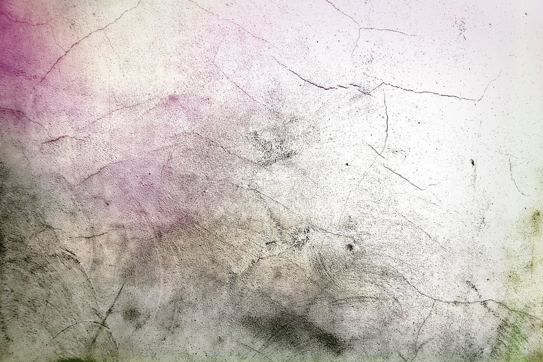 Cracked concrete wall surface with faded pink and green watercolor stains and intricate weathered fissures.