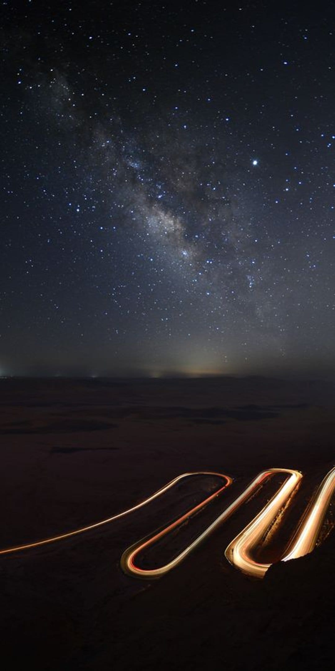 Winding road light trails glow beneath a star-filled Milky Way galaxy in a vertical night sky composition.