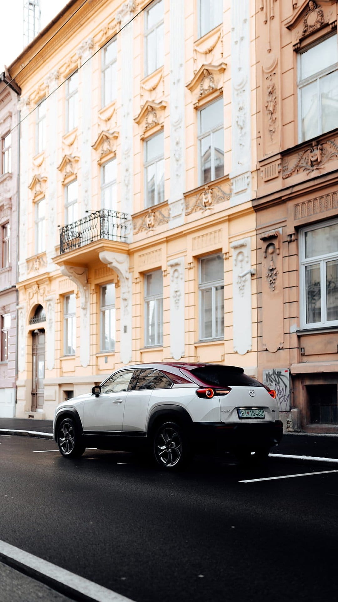 White Mazda CX-30 with a red roof parked on a street in front of a cream-colored ornate European building.