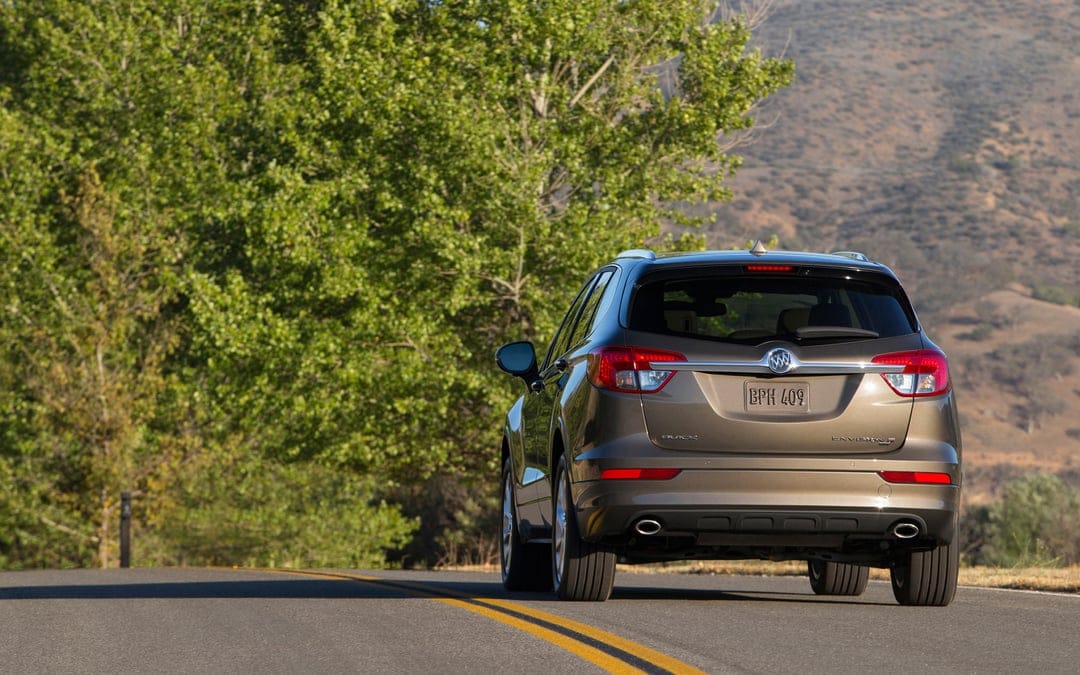 Rear view of a bronze Buick SUV driving on a curved road flanked by green trees and distant sunlit hills.