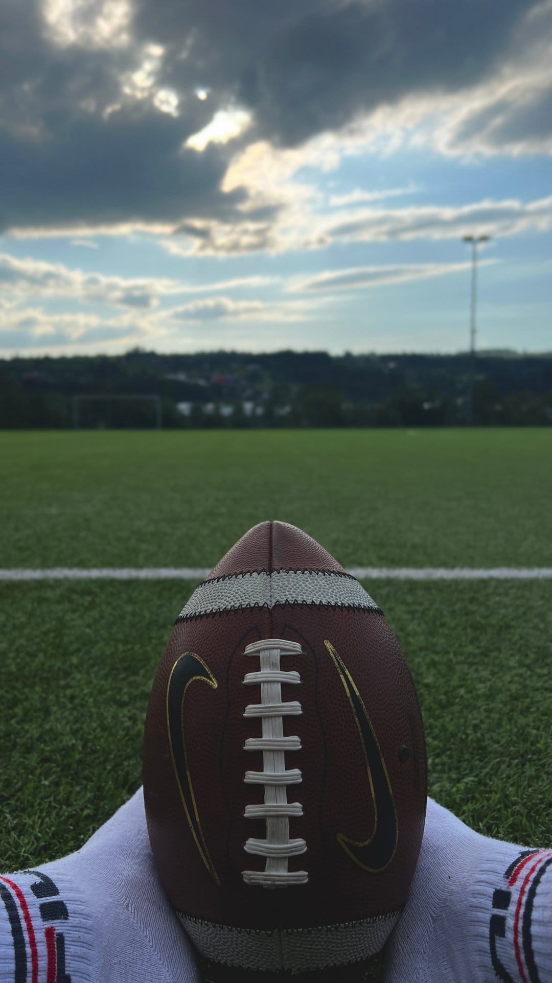 American football with white laces rests on green artificial turf under a dramatic sky with breaking sunlight.