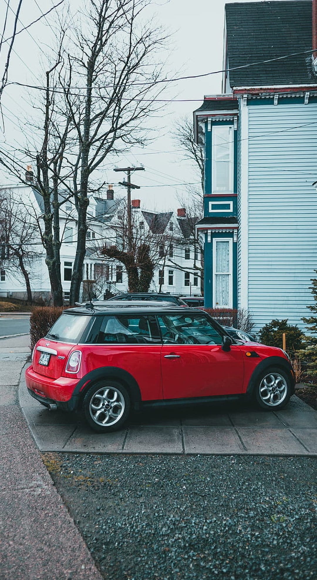 Red Mini Cooper parked on a grey tiled driveway in front of a blue suburban house with bare tree branches.