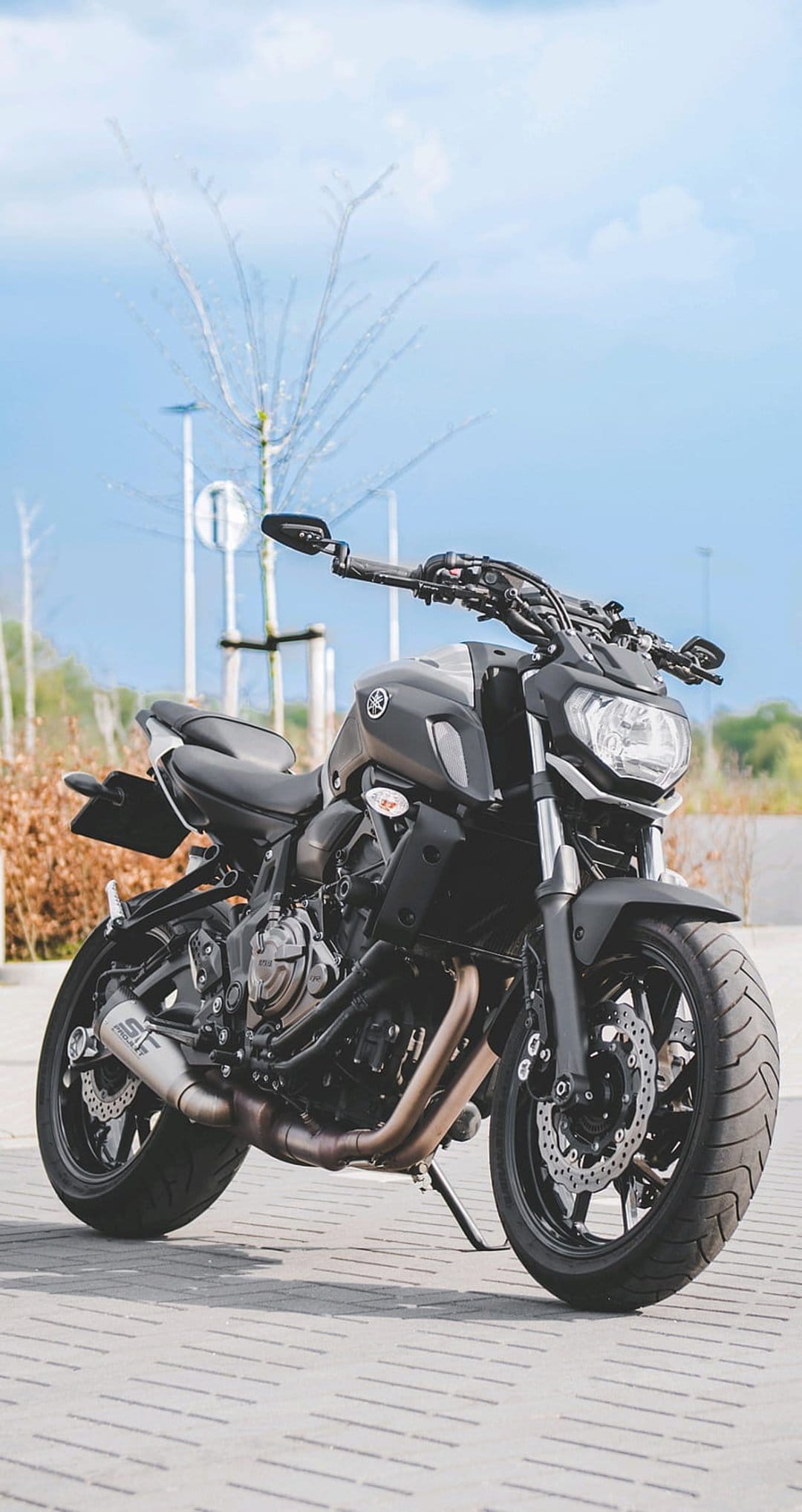 Yamaha MT-07 motorcycle in matte black parked on brick pavement under a blue sky with soft trees in background.