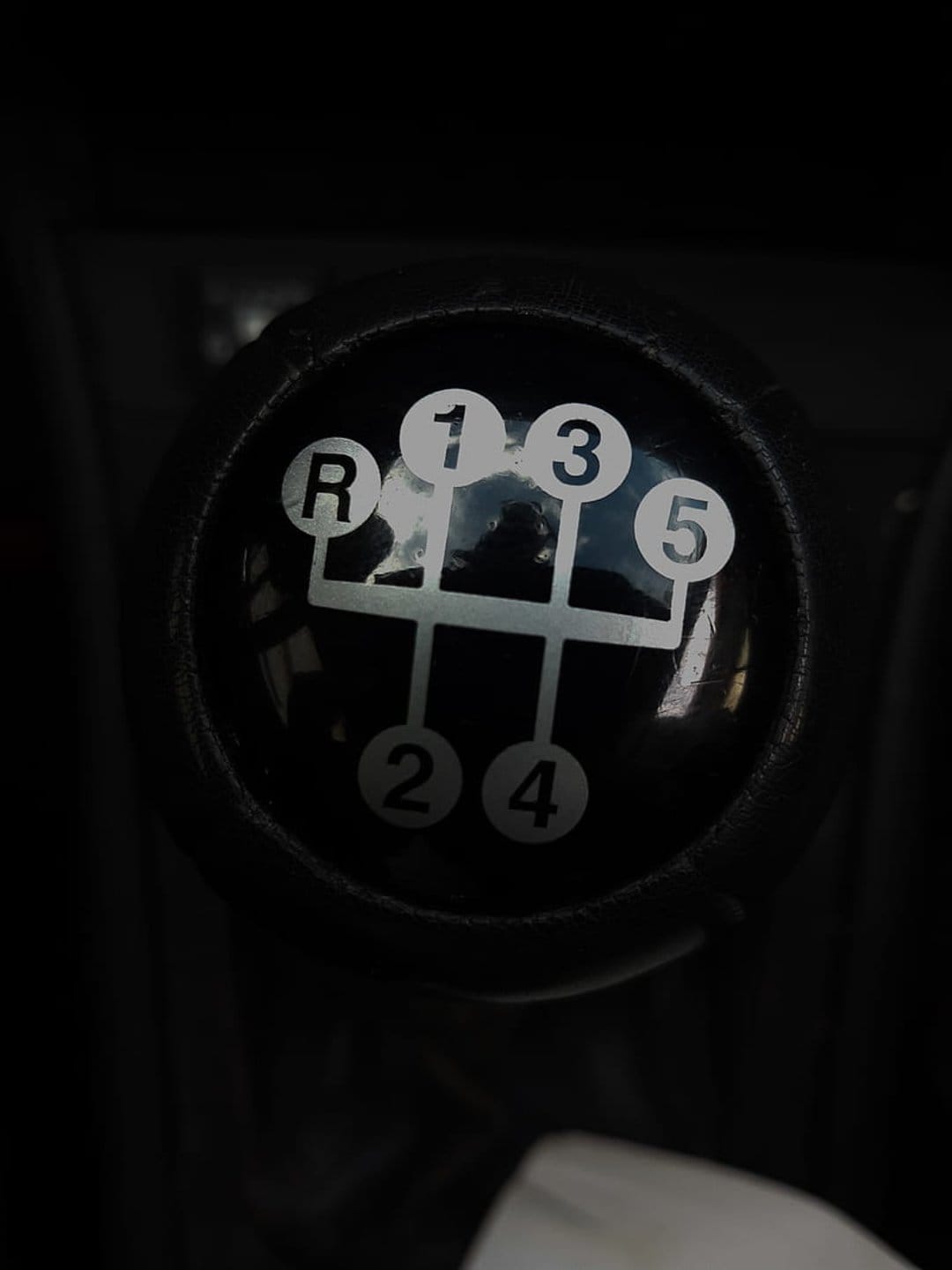 Black 5-speed manual gear shift knob with white H-pattern lettering reflects a dim car interior environment.