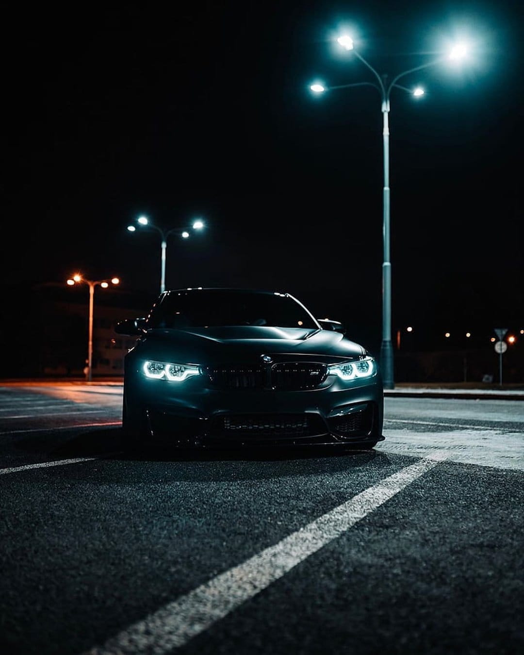Black BMW M sedan parked on dark asphalt under warm streetlights with glowing LED headlights at night.