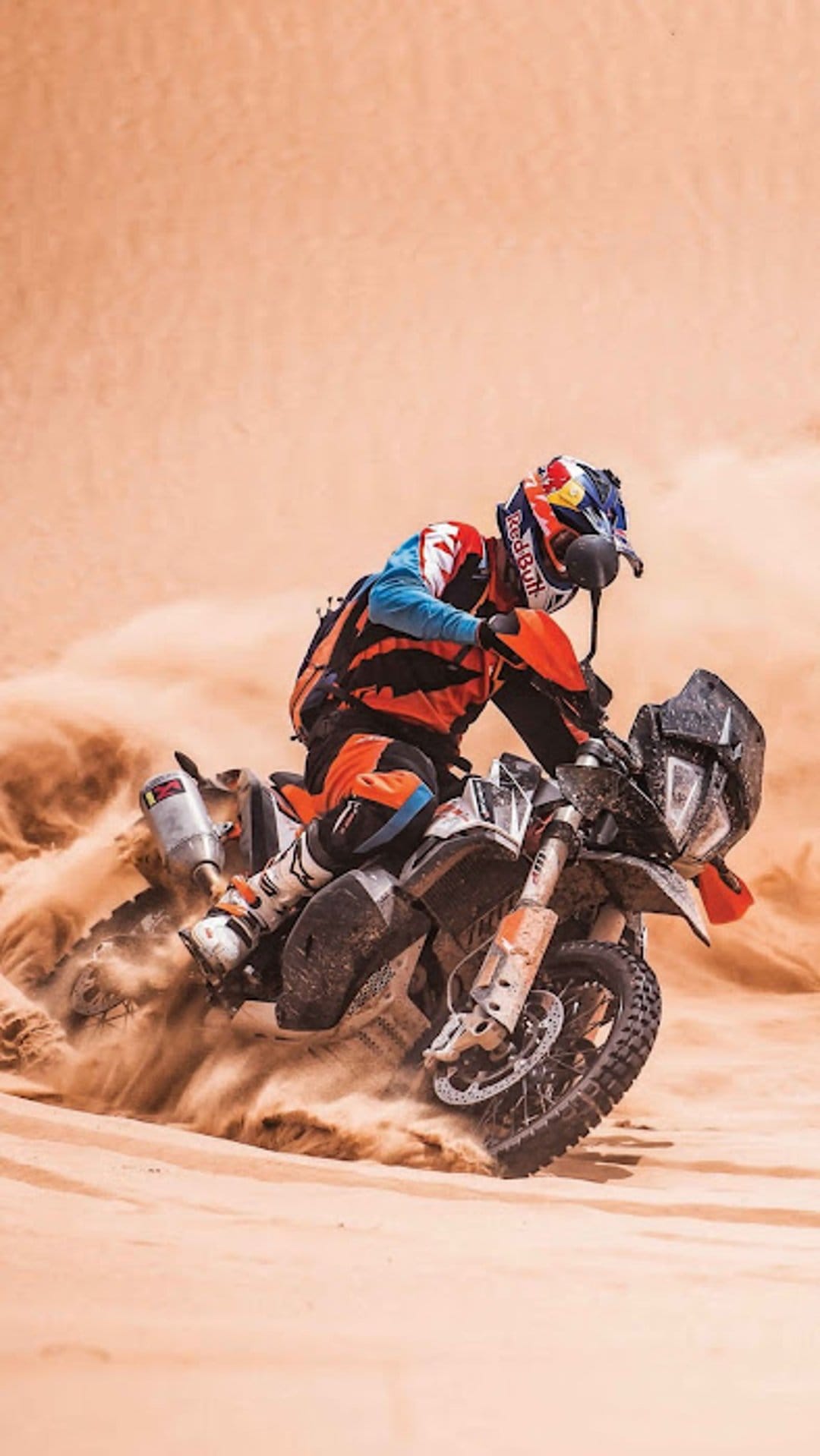 Motocross rider in orange gear leans into a steep desert dune, spraying a massive cloud of golden sand.