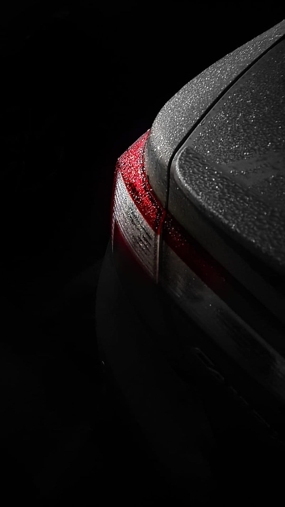 Dark car tail light covered in glistening raindrops with dramatic low-light reflections on a wet vehicle surface.