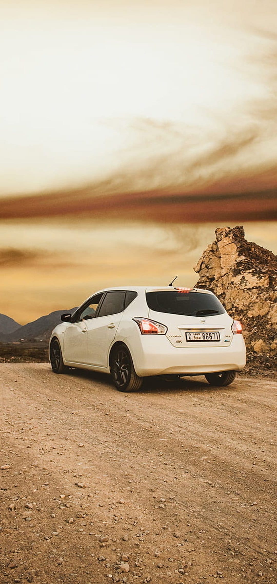 White Nissan hatchback on a dirt road with a dramatic fiery orange sunset sky and distant desert mountains.
