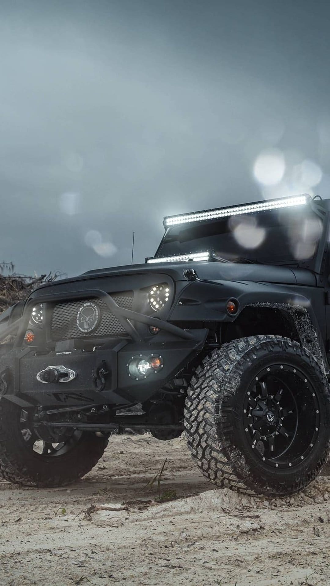 Black Jeep Wrangler with aggressive tires and LED light bars parked on sand under a cloudy, atmospheric sky.