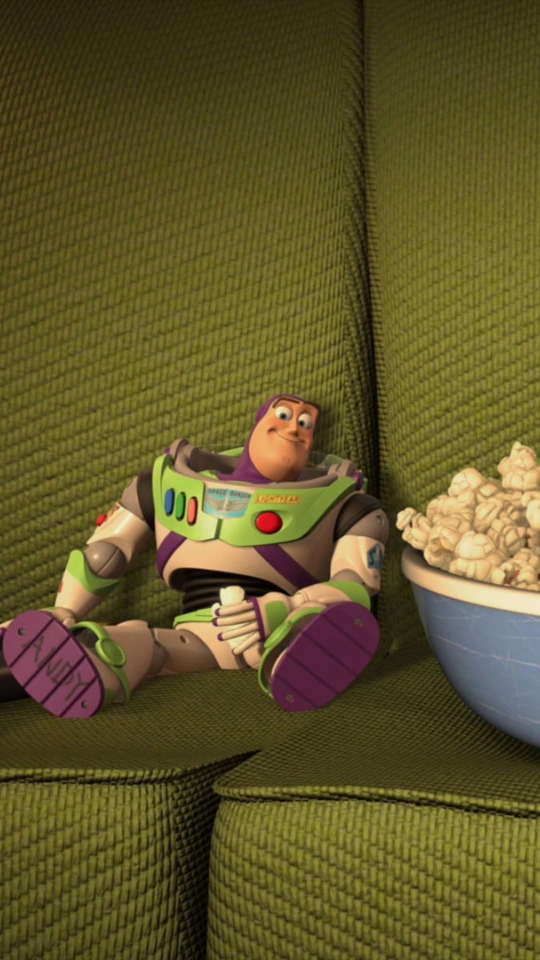 Buzz Lightyear sits on a green textured couch with a white bowl of popcorn beside his armored space suit legs.
