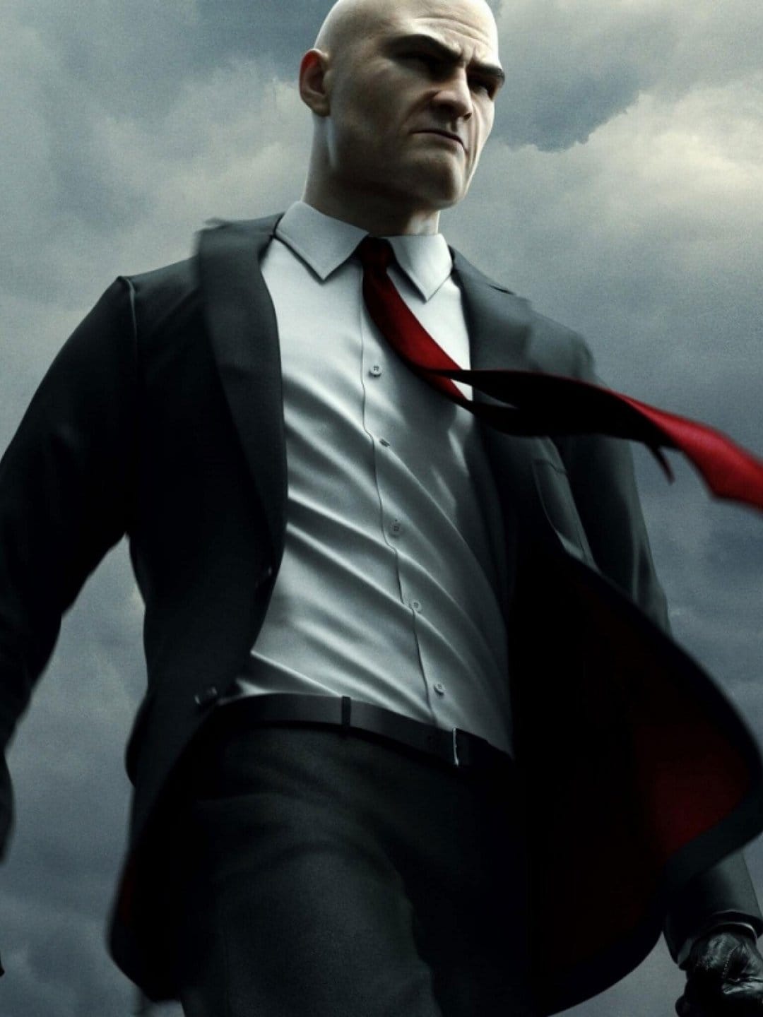 Bald man in a black suit and red tie standing against a dark stormy sky with wind blowing his clothing.