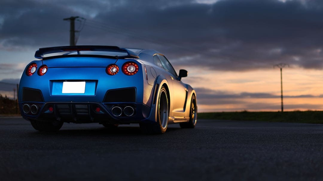 Rear view of a blue widebody Nissan GT-R R35 on an asphalt road at sunset with glowing circular taillights.
