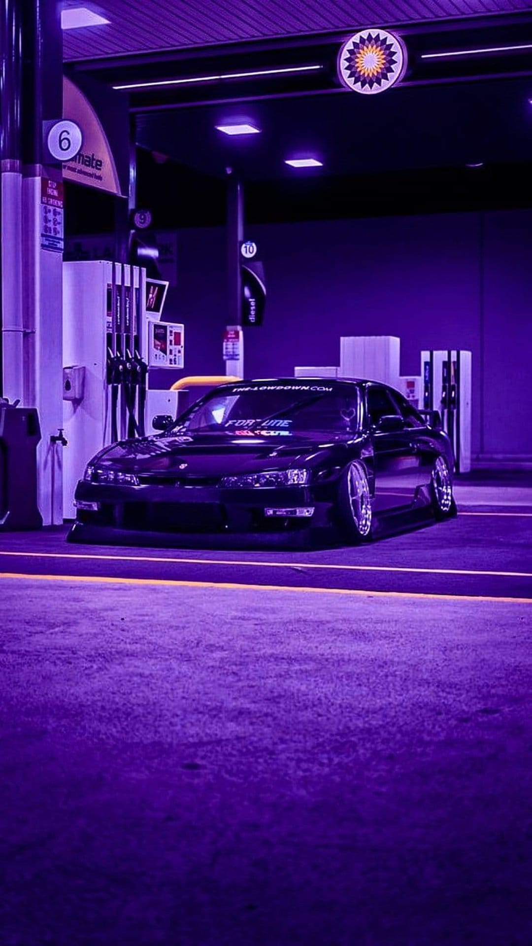 Black sports car parked under purple neon lights at a gas station with reflective surfaces and fuel pumps.
