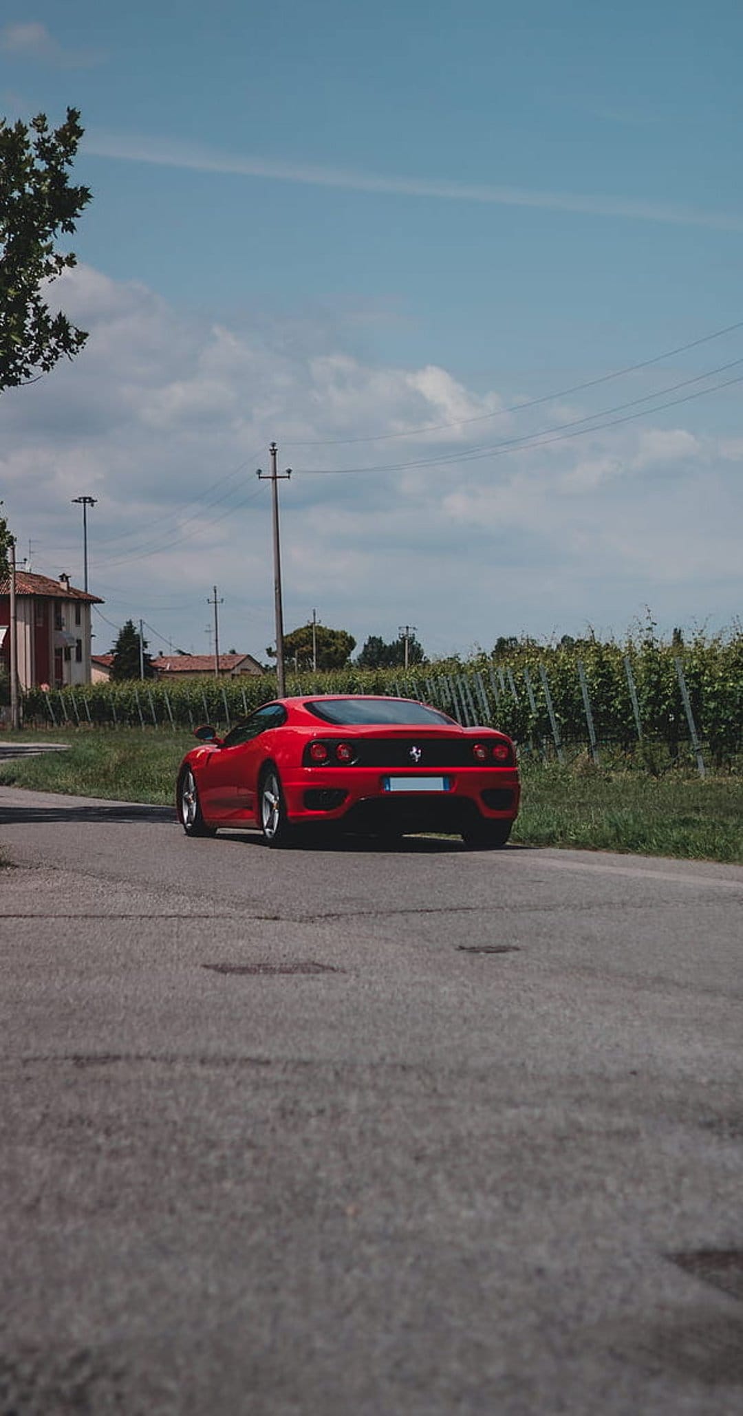 Red Ferrari sports car driving on a road past green vineyards and old Italian houses under a blue sky.