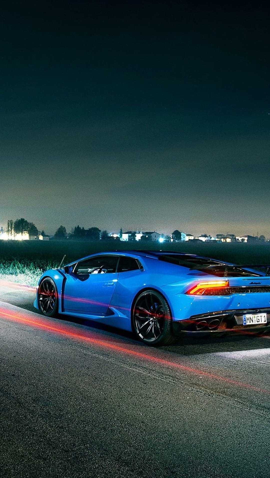 Blue Lamborghini Huracan speeds on dark asphalt leaving glowing red light trails beneath blurred nighttime trees.