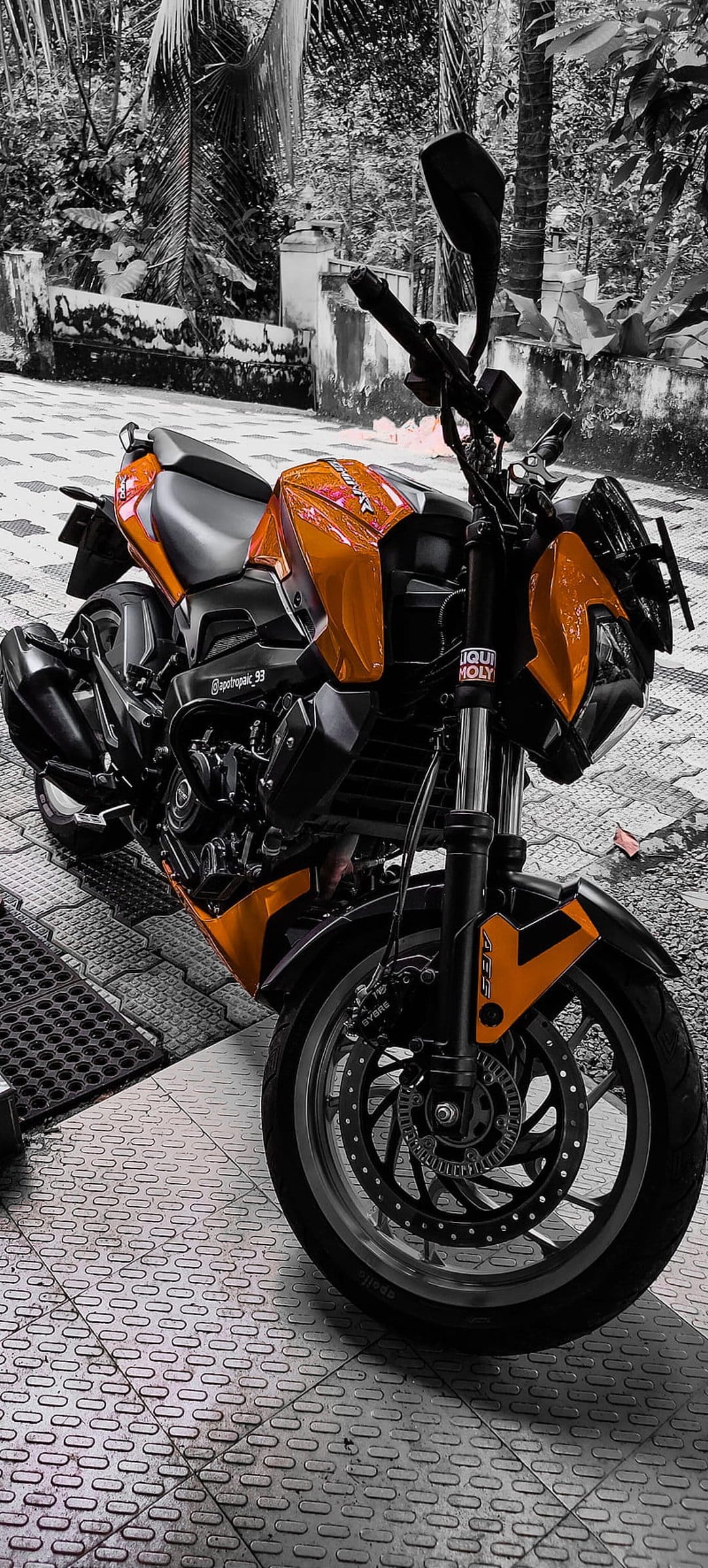 Orange sportbike parked on textured grey pavement showing mechanical details like front suspension and brakes.