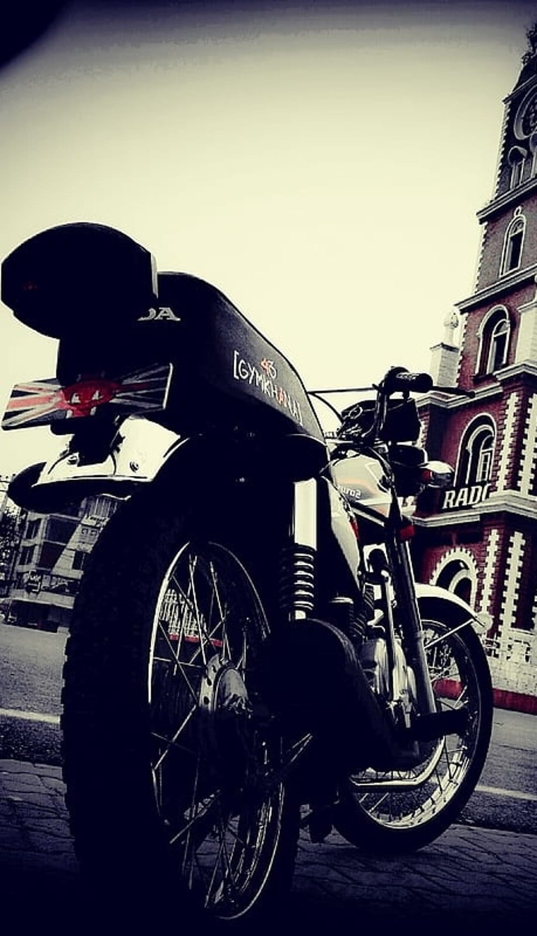 Vintage motorcycle with a Union Jack fender sits on a cobblestone street near a red brick clock tower.