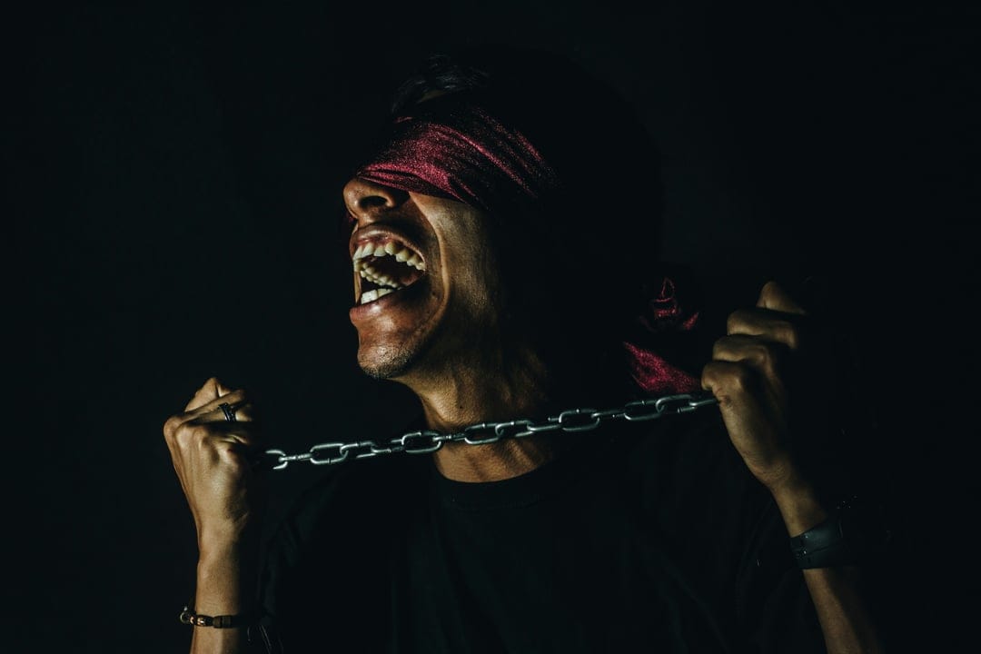 Man screaming with a red blindfold while gripping heavy iron chains against a dark, dramatic background.