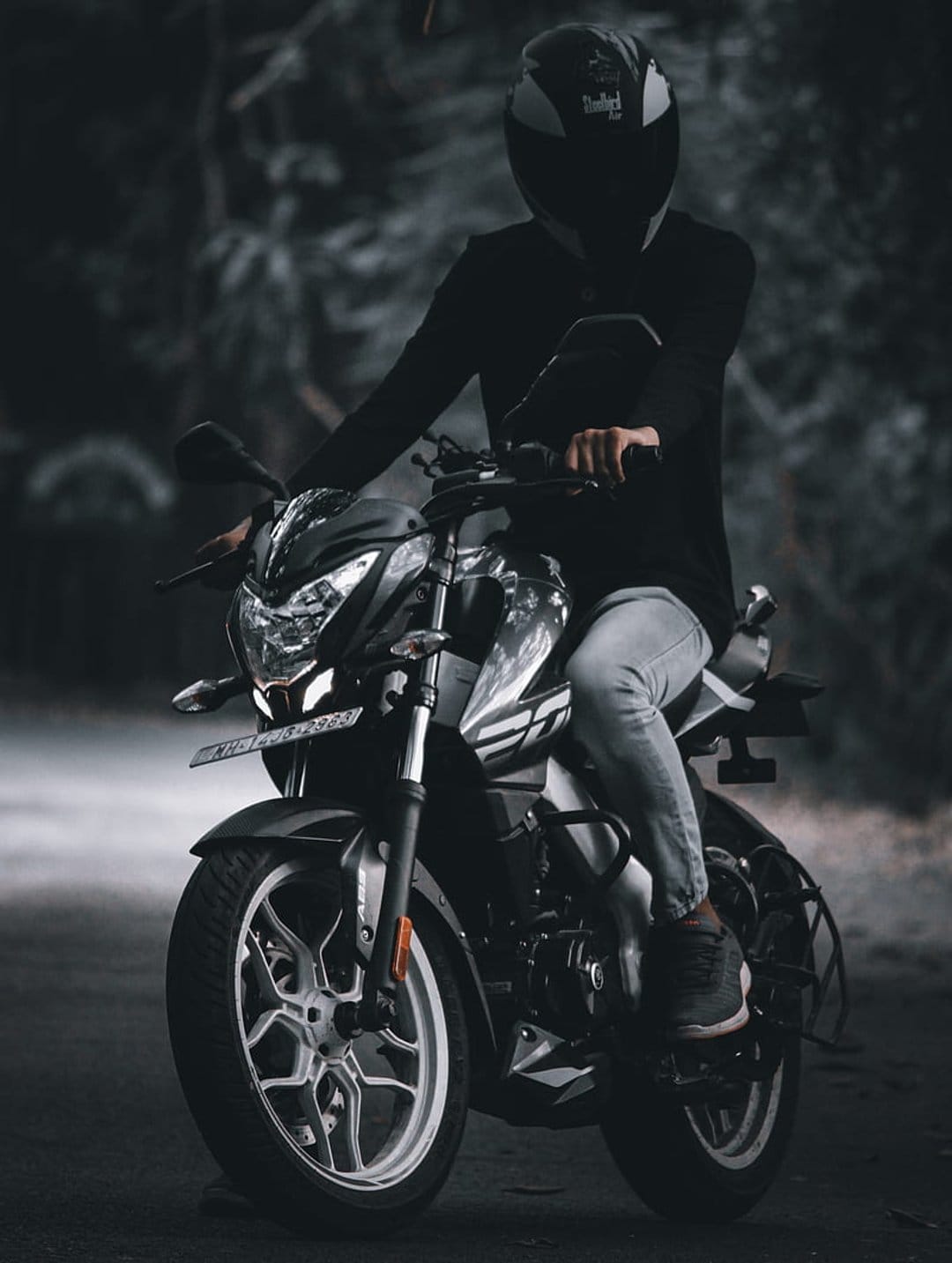 Biker in dark gear sits on a motorcycle on a paved road surrounded by a blurred, moody forest at dusk.