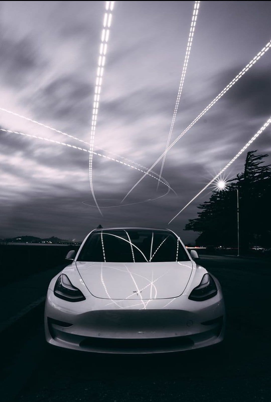 White Tesla car parked on a dark road under a cloudy sky filled with glowing neon light trails at twilight.