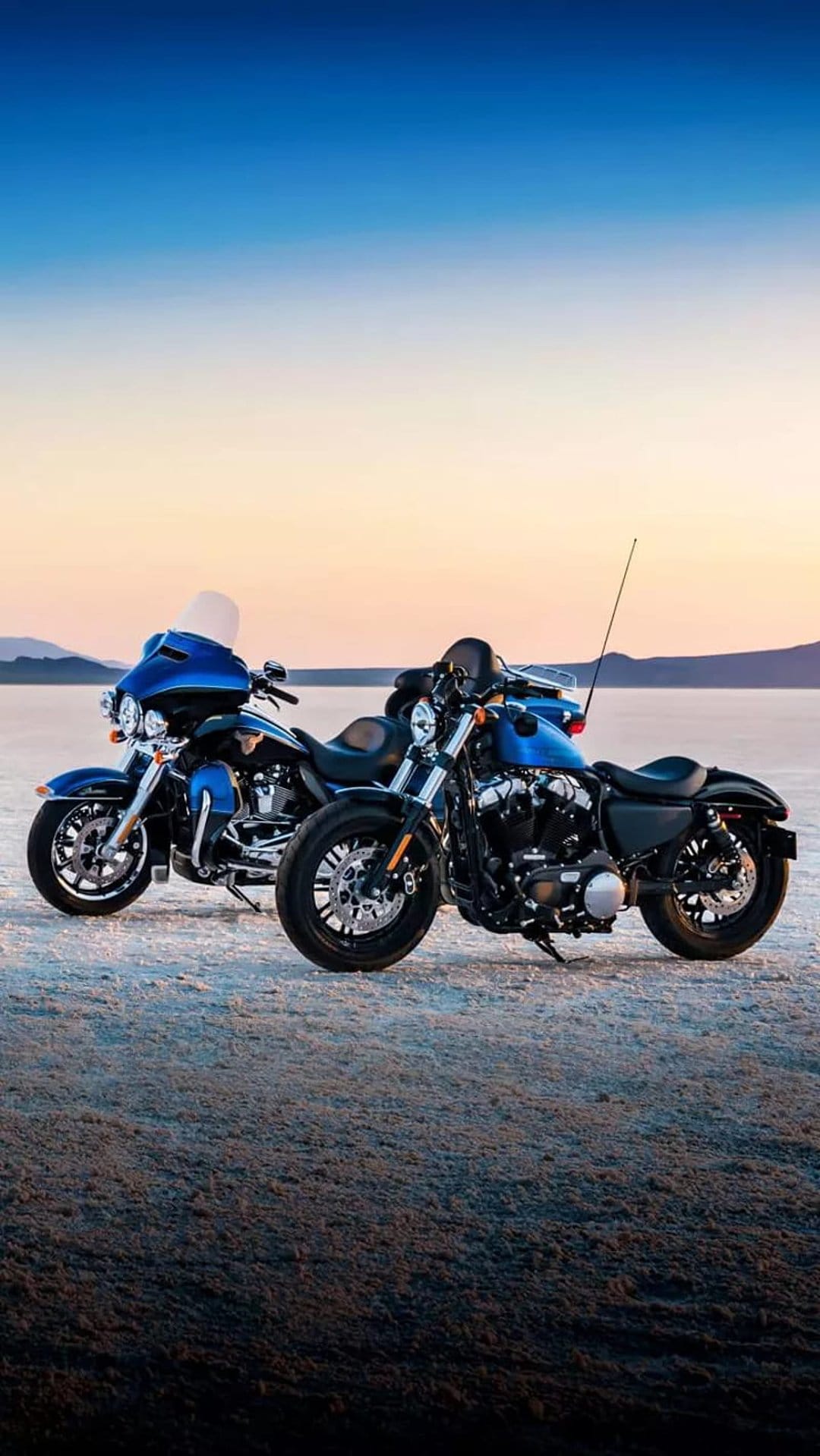 Twin blue Harley-Davidson motorcycles parked on a sandy beach under a gradient orange and blue sunset sky.