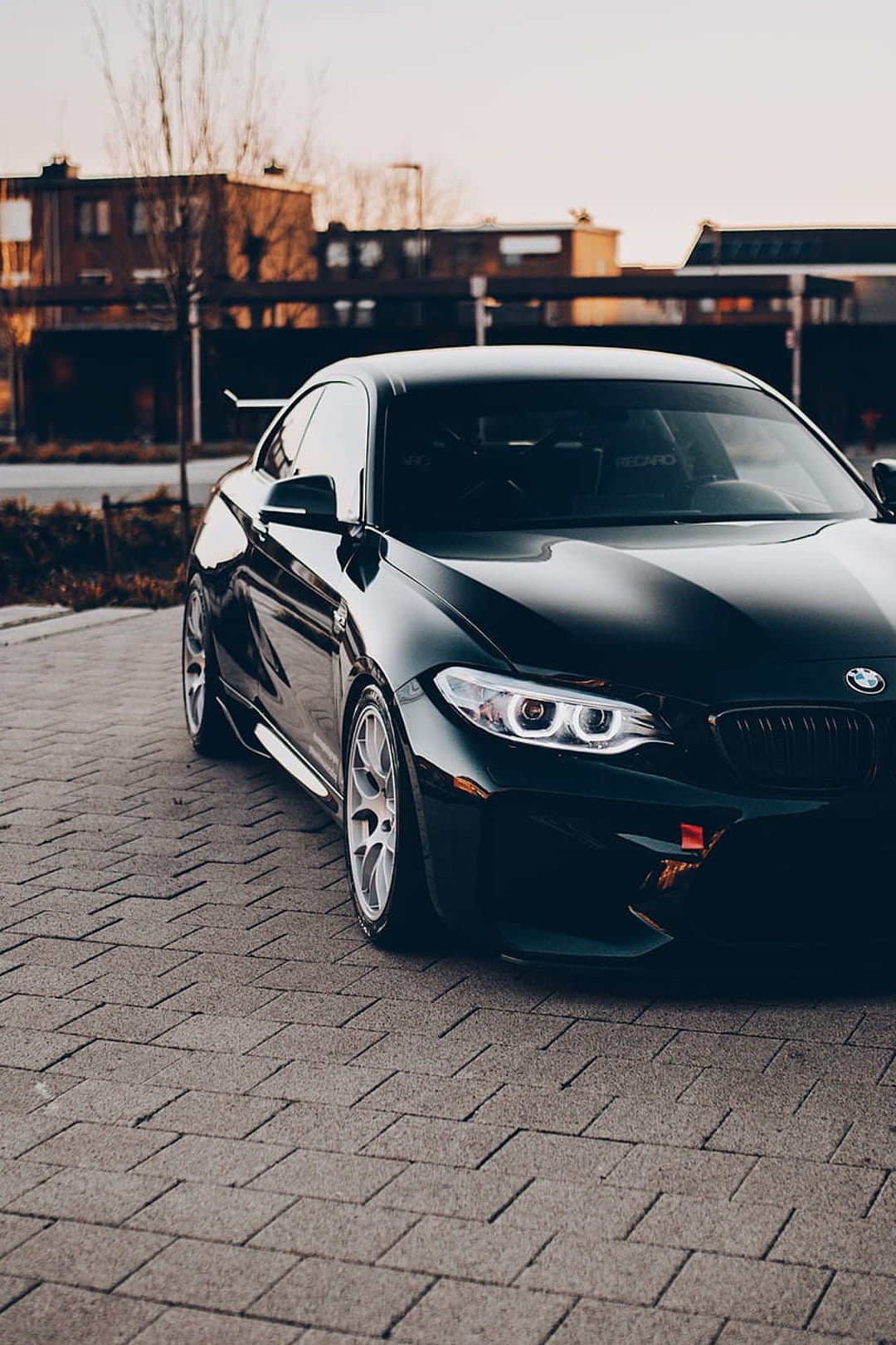 Black BMW M2 coupe on a cobblestone street at sunset with glowing headlights and a blurred urban background.
